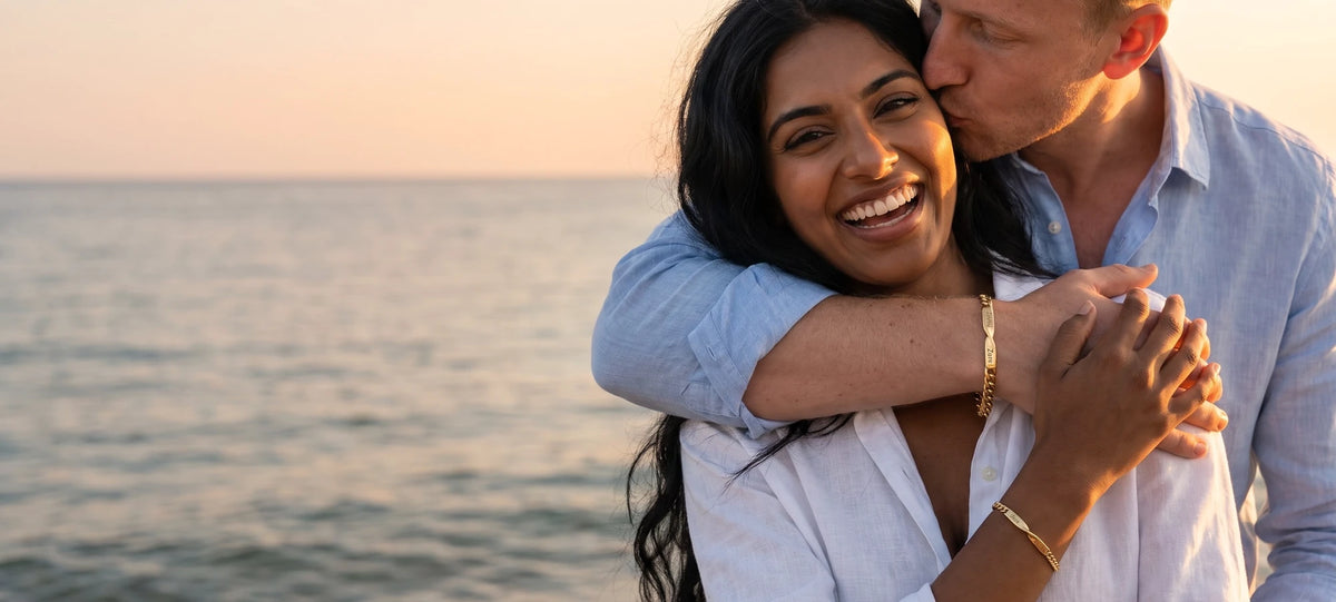 Ein Paar lacht am Strand bei Sonnenuntergang, beide tragen ihr passendes Armband in Gold